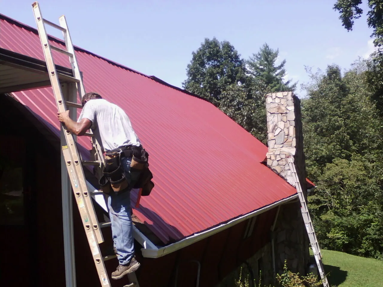 Roofer on a ladder beside a red metal roof and stone chimney — Soffit & Fascia Repair by Roofing Brothers in Lindale